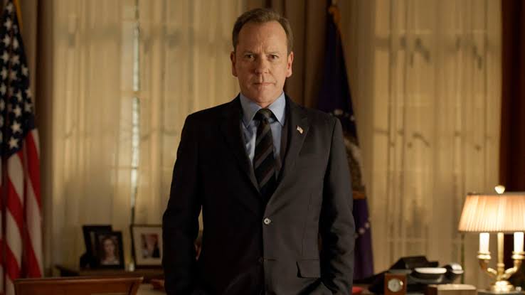 Tom Kirkman (Kiefer Sutherland) de terno escuro, em pé num escritório com a bandeira dos Estados Unidos ao fundo