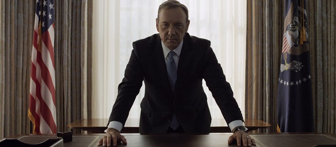Frank Underwood (Kevin Spacey) apoiado numa escrivaninha, olhando sério pra câmera, com a bandeira dos Estados Unidos e o brasão presidencial atrás dele