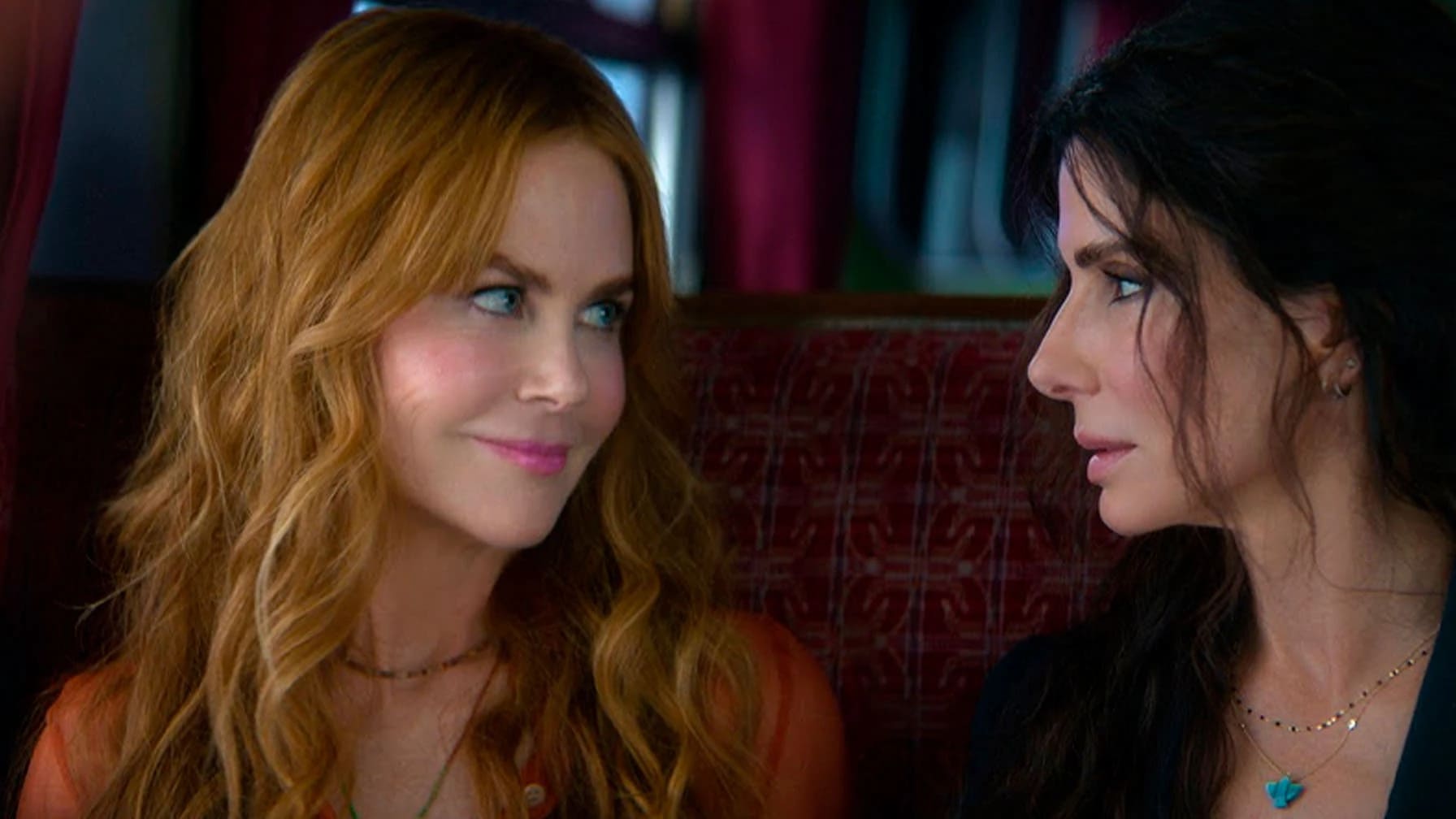Imagem de trecho do filme Da Magia à Sedução: Feitiço de Amor (2026) com Sally (Sandra Bullock), desconfiada, e Gillian Owens (Nicole Kidman), com um sorriso, olhando uma pra outra enquanto estão sentadas no banco de um trem.
