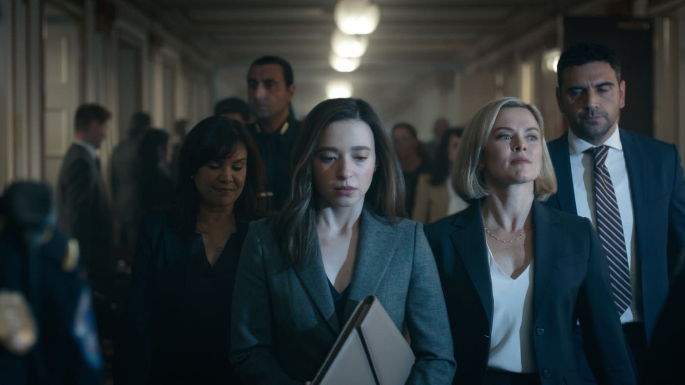 Cena do filme O Outro Lado das Redes, com Frances Haugen (Mikey Madison) caminhando por um corredor ao lado de uma mulher loira e de outros profissionais, todos vestidos formalmente.