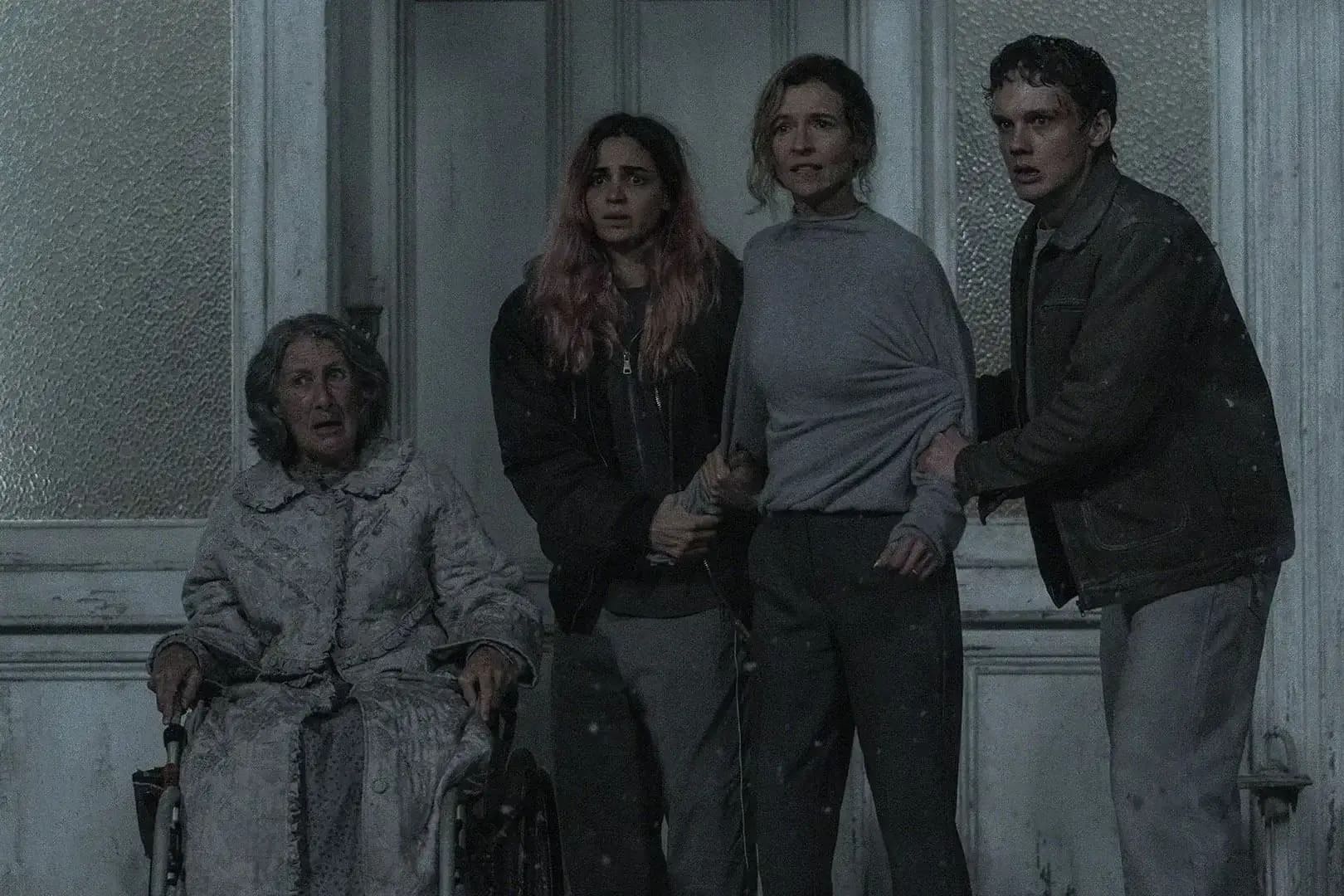 Cena do filme A Morte do Demônio: Em Chamas, com os personagens Polly (Maude Davey), de cadeira de rodas, Alice (Souheila Yacoub), Susan (Tandi Wright) e Joseph (Hunter Doohan), em pé ao lado de Polly, estão assustados em frente a porta de casa enquanto observam algo fora do enquadramento.