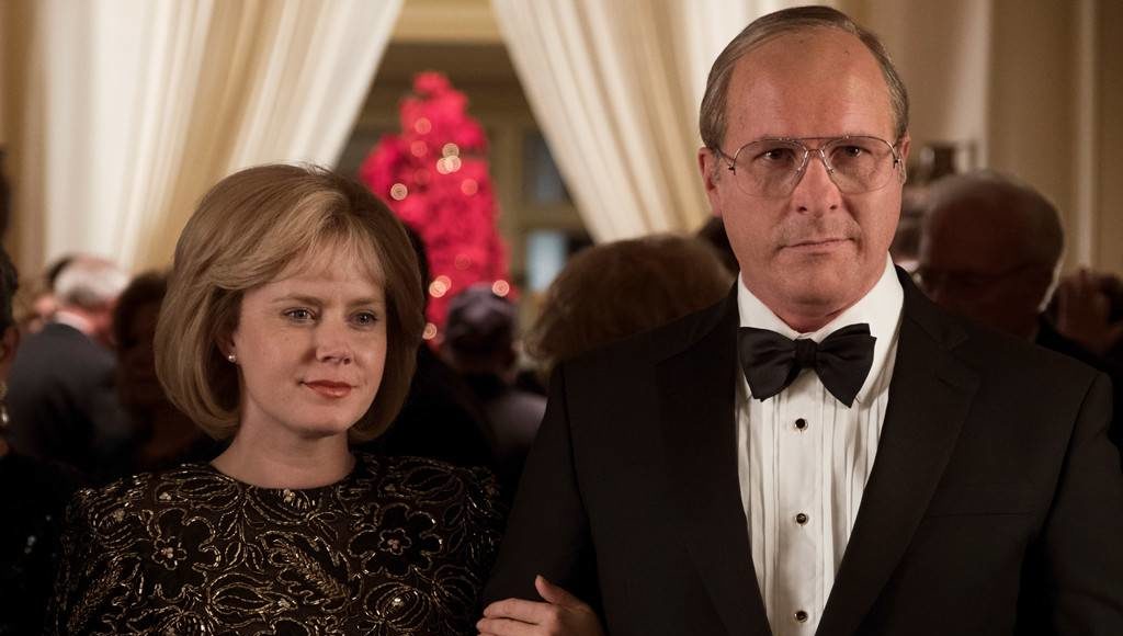 Lynne Cheney (Amy Adams) e Dick Cheney (Christian Bale) em um evento formal, cercados por convidados, com uma árvore de Natal decorada ao fundo