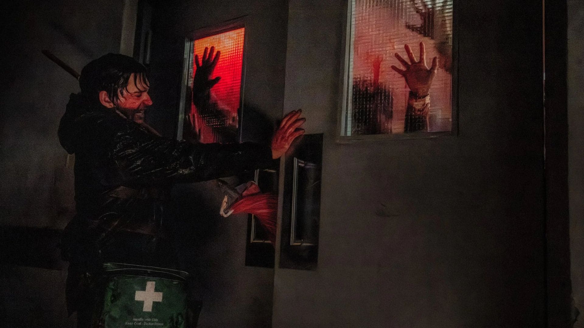 Bryan (Austin Abrams) ensanguentado bloqueando a porta de uma sala repleta de zumbis com um esfregão de madeira no filme Resident Evil (2026). Pelo vidro da porta, é possível ver as mãos ensanguentadas dos infectados trancados na sala.