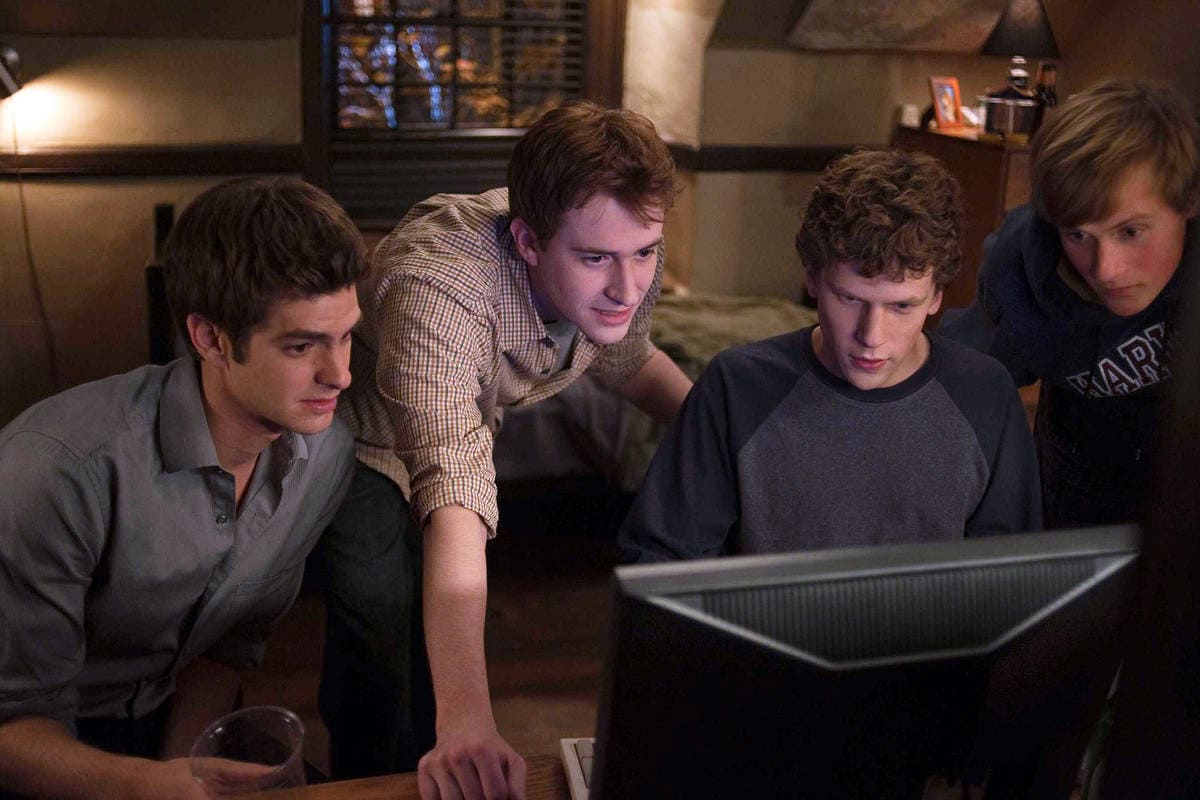 Cena do filme A Rede Social, com Mark Zuckerberg (Jesse Eisenberg), Eduardo Saverin (Andrew Garfield) e outros dois estudantes reunidos em frente a um computador, observando a tela enquanto desenvolvem a plataforma.