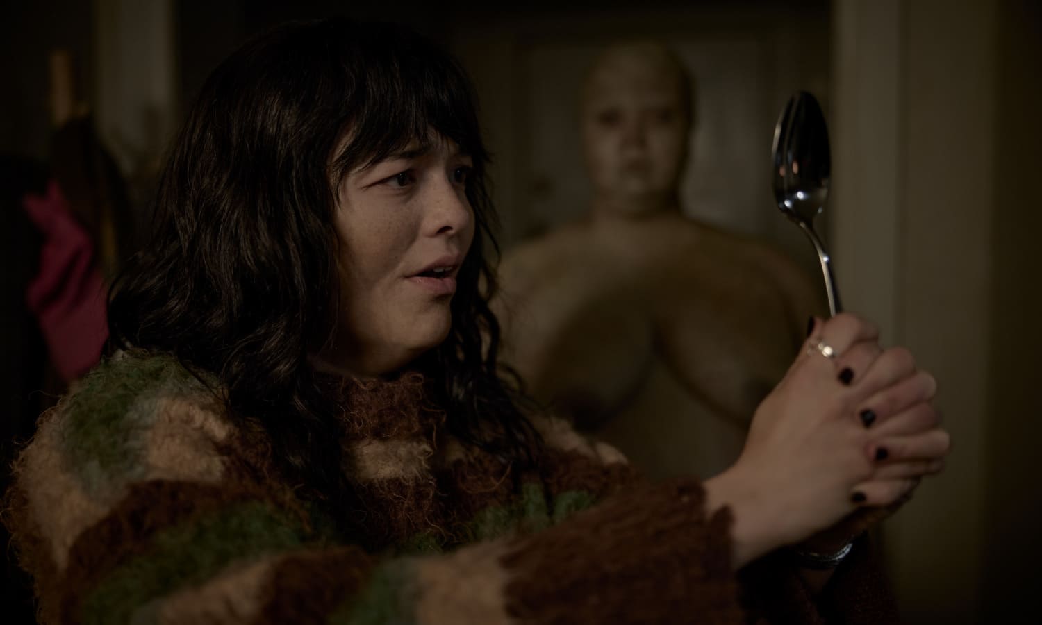 Hana (Midori Francis) segura uma colher diante de seu rosto enquanto uma figura humana de aparência fantasmagórica aparece ao fundo, em uma cena sombria do filme de terror Insaciável.