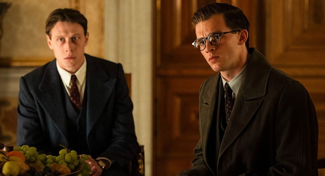 Paul von Hartmann (Jannis Niewöhner) e Hugh Legat (George MacKay) sentados a uma mesa com uma travessa de frutas, numa sala decorada em madeira