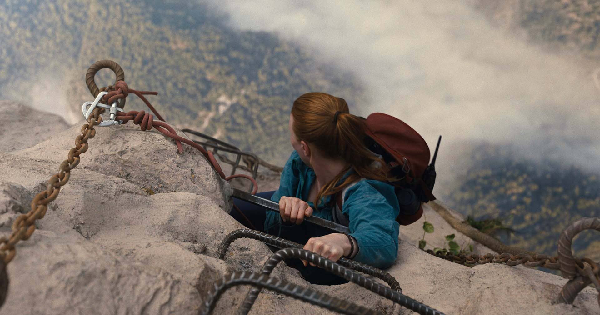 Cena do filme A Queda 2: No Limite, com Jax Hunter (Harriet Slater), de mochila nas costas, escalando por uma escada de metal cravada no penhasco e olhando pra baixo.
