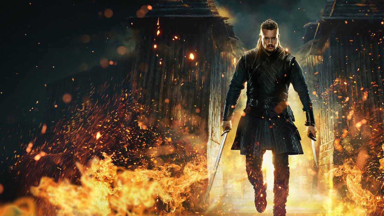 Imagem promocional de O Último Reino, mostrando o protagonista Uhtred (Alexander Dreymon) em vestes de batalha medieval caminhando em meio a uma construção em chamas, segurando uma espada em cada mão.