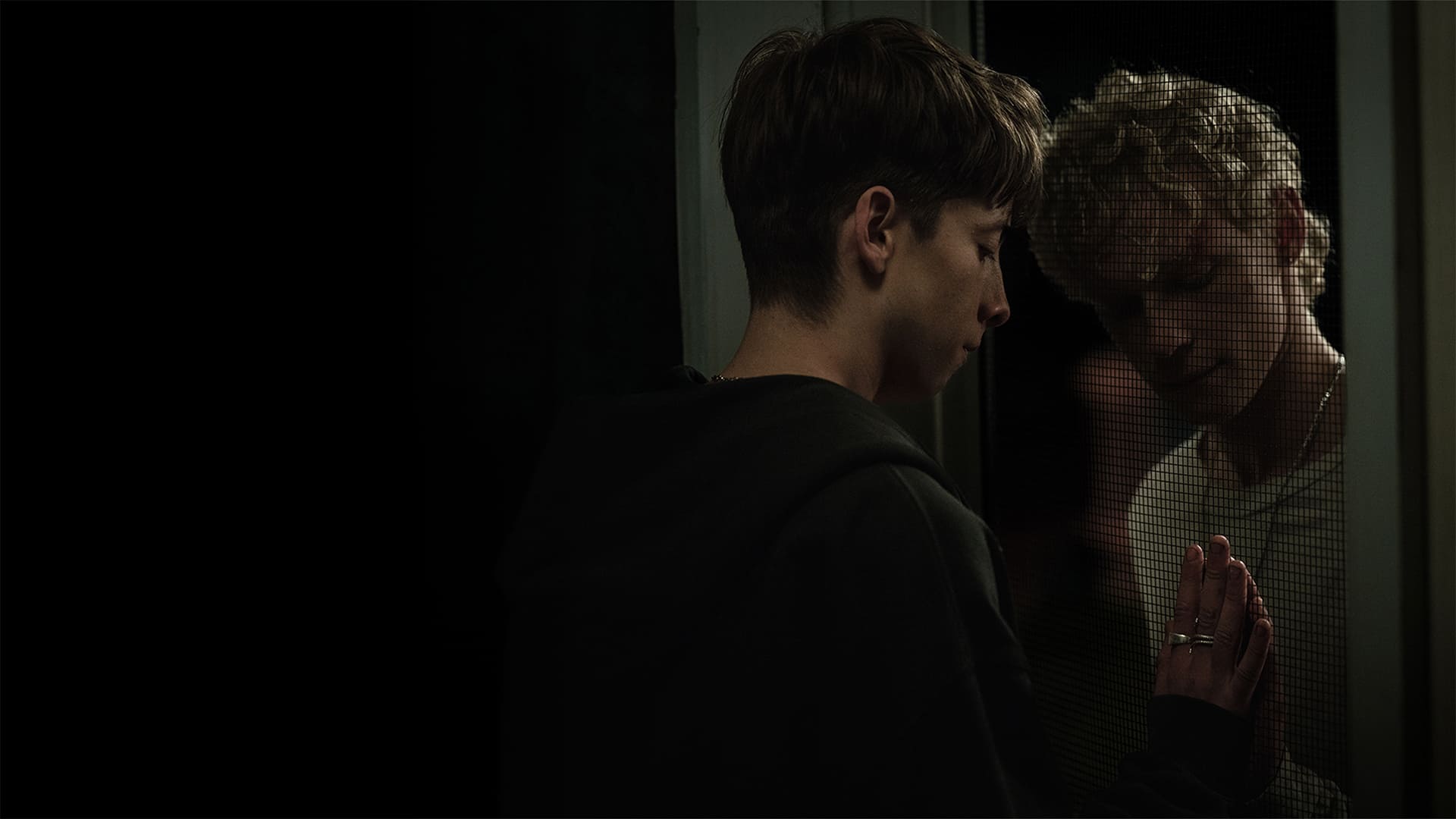 Niam (Joe Bird) e Ryan (Stacy Clausen) encostando as mãos através de uma porta telada em cena do filme Leviticus.