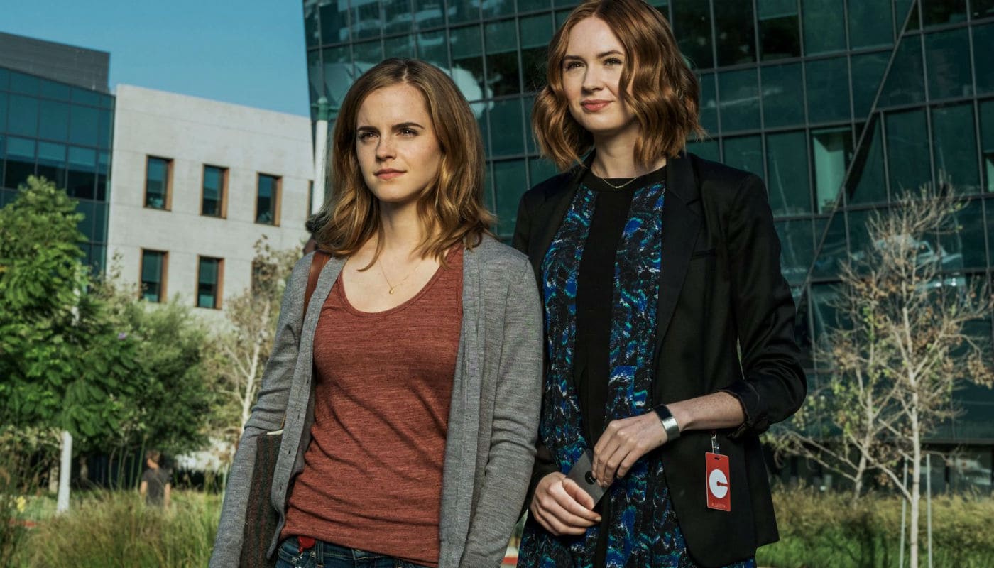 Cena do filme O Círculo, com Mae Holland (Emma Watson) e Annie Allerton (Karen Gillan) em frente a um prédio moderno de fachada envidraçada.