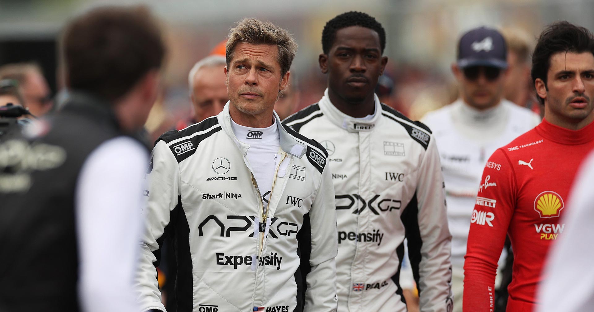 F1: Sonny Hayes (Brad Pitt) e Joshua Pearce (Damson Idris) de macacão de corrida, caminhando pelo grid ao lado de outros pilotos
