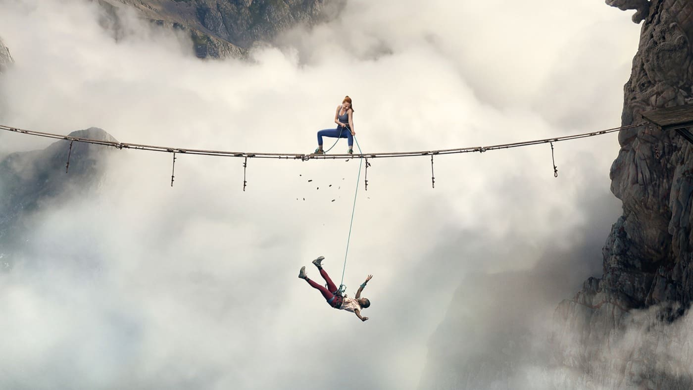Imagem promocional do filme A Queda 2: No Limite, com Luce (Arsema Thomas) suspensa por uma corda abaixo de uma ponte estreita de madeira, enquanto Jax (Harriet Slater) tenta ajudá-la segurando a corda. Ambas estão em meio a montanhas e nuvens.